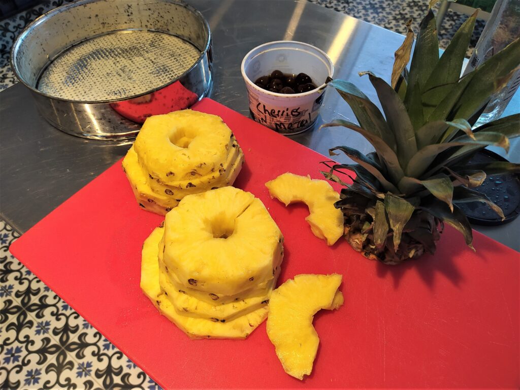 Pineapple upside down cut preparation with greased pan, cut pineapples and cherries.